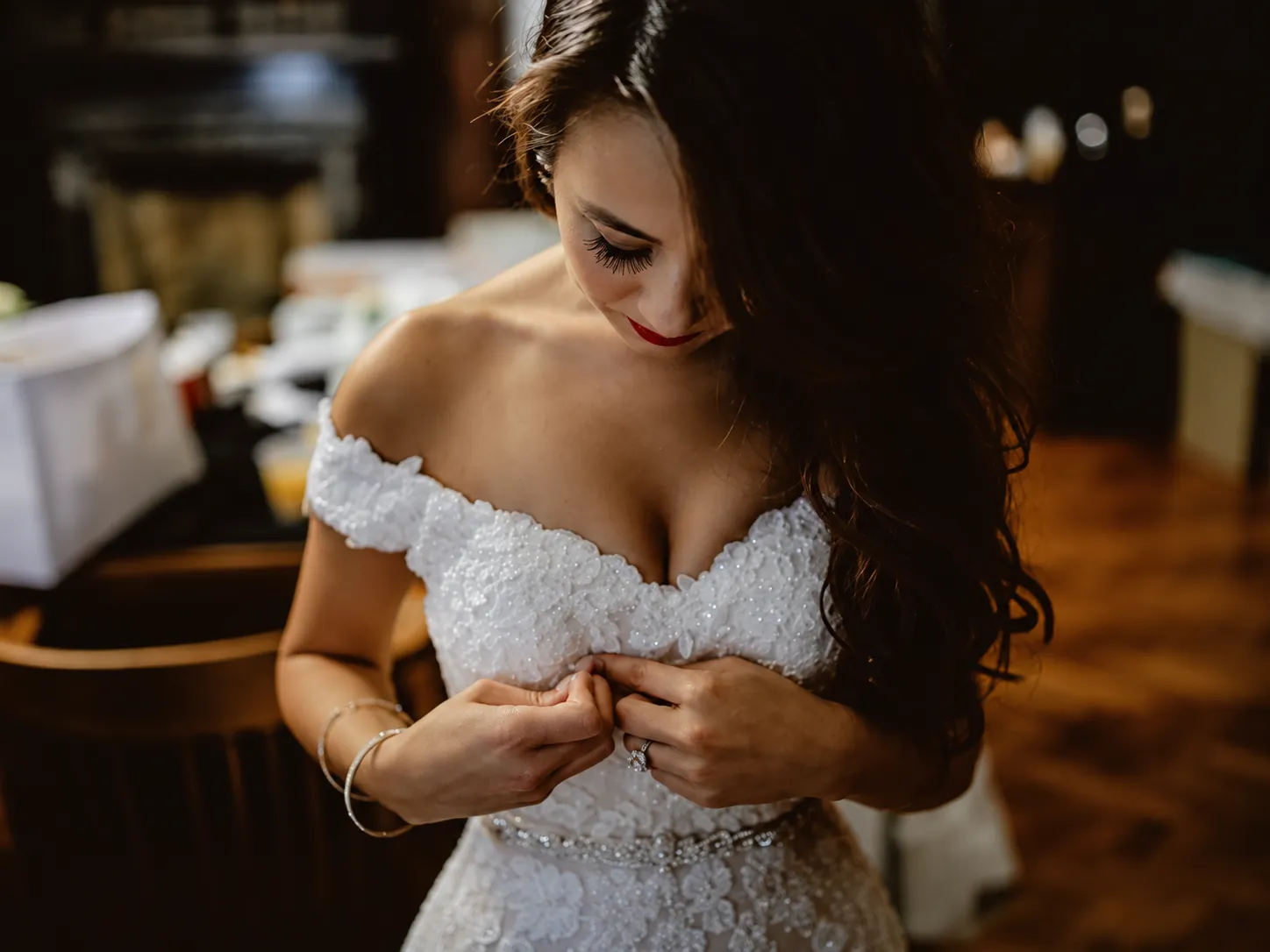 A bride in an off-the-shoulder lace gown, looking down as she fastens her dress